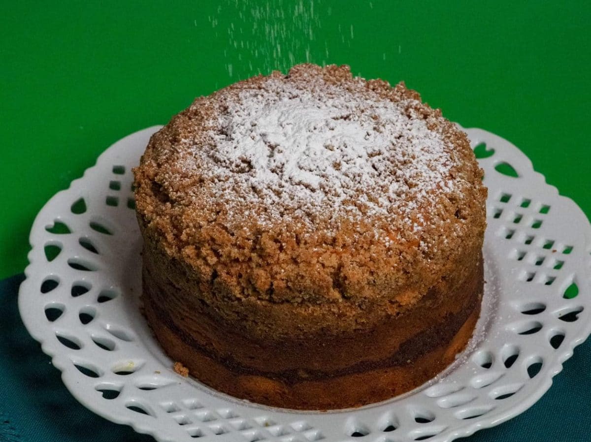 Coffee cake on a white decorated cake plate with a rain of icing sugar on top of a green placemat.