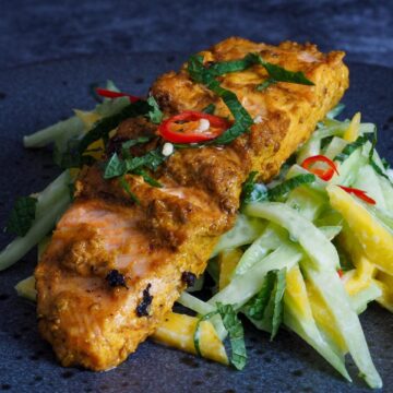 Grilled salmon placed on top of mango and cucumber salad garnished with chilli slices and shredded fresh on a dark grey plate. mint
