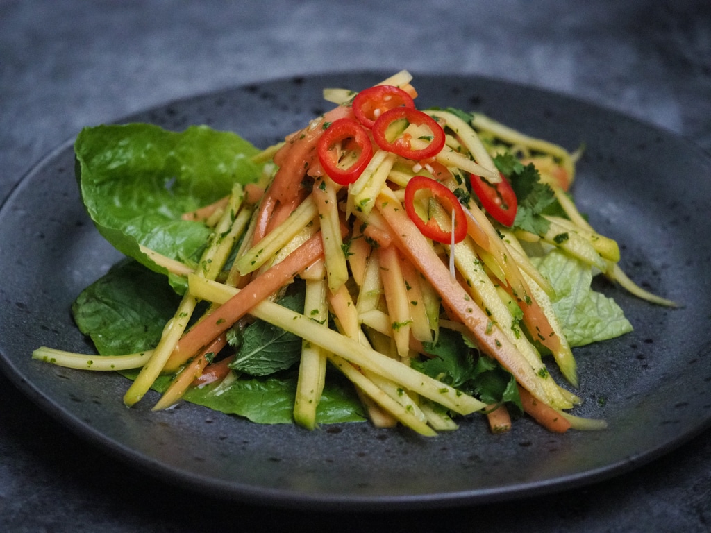 Slivers Mango, papaya and romaine lettuce garnished with red chilli slices on a black plate.