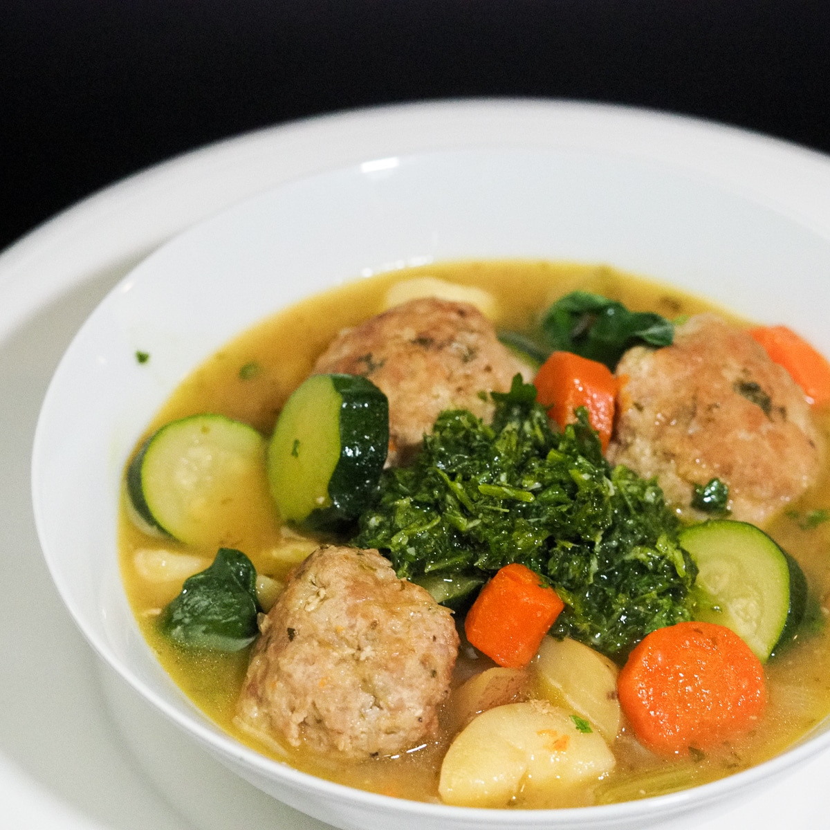 Meatballs. chopped carrots, potatoes, zucchini with broth in a white bowl topped with green pesto