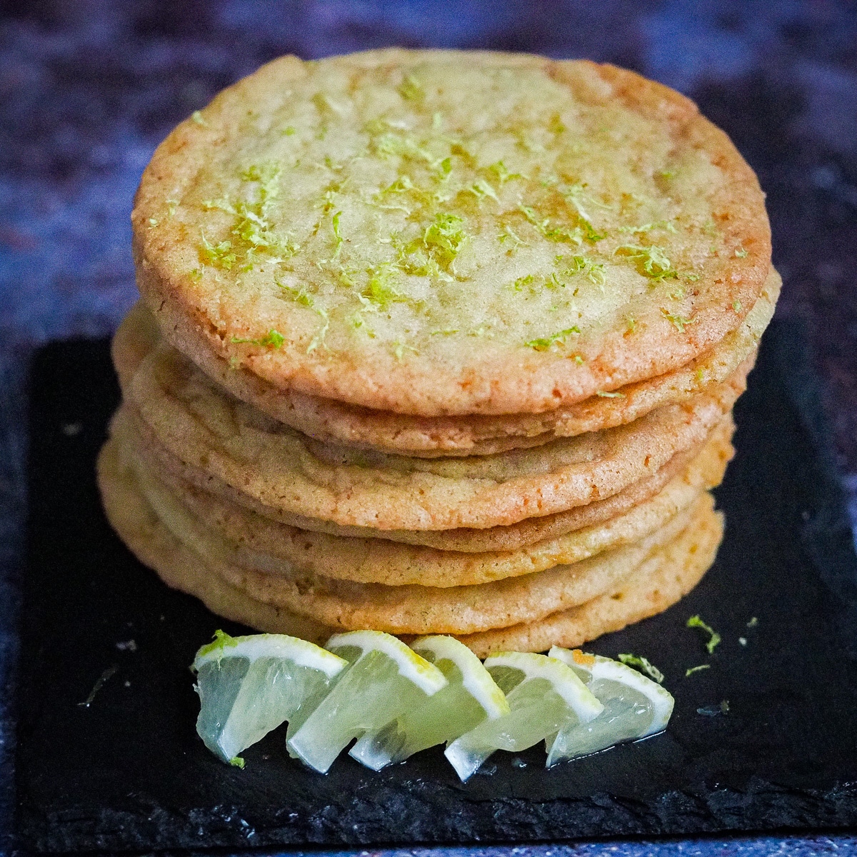 The best thin and crispy lemon cookies garnished with lemon zest stacked on a black serving dish with thin quarter slices of fresh lemons on the side.