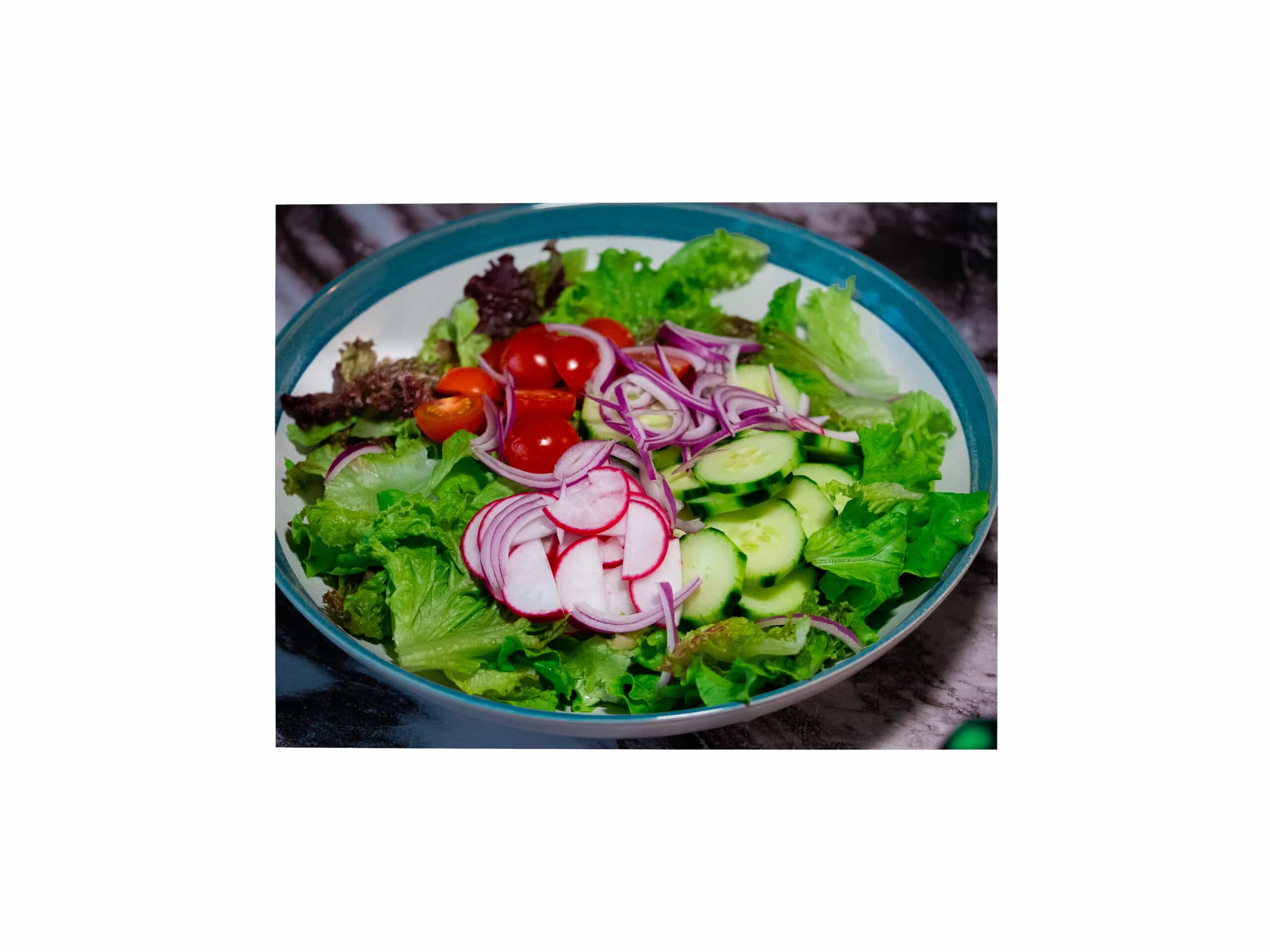 A big bowl of mixed chopped lettuce, sliced red radish, sliced cucumber,Sliced cherry tomatoes and thinly sliced red onion with lemon and ginger vinaigrette on a large blue and white serving bowl.