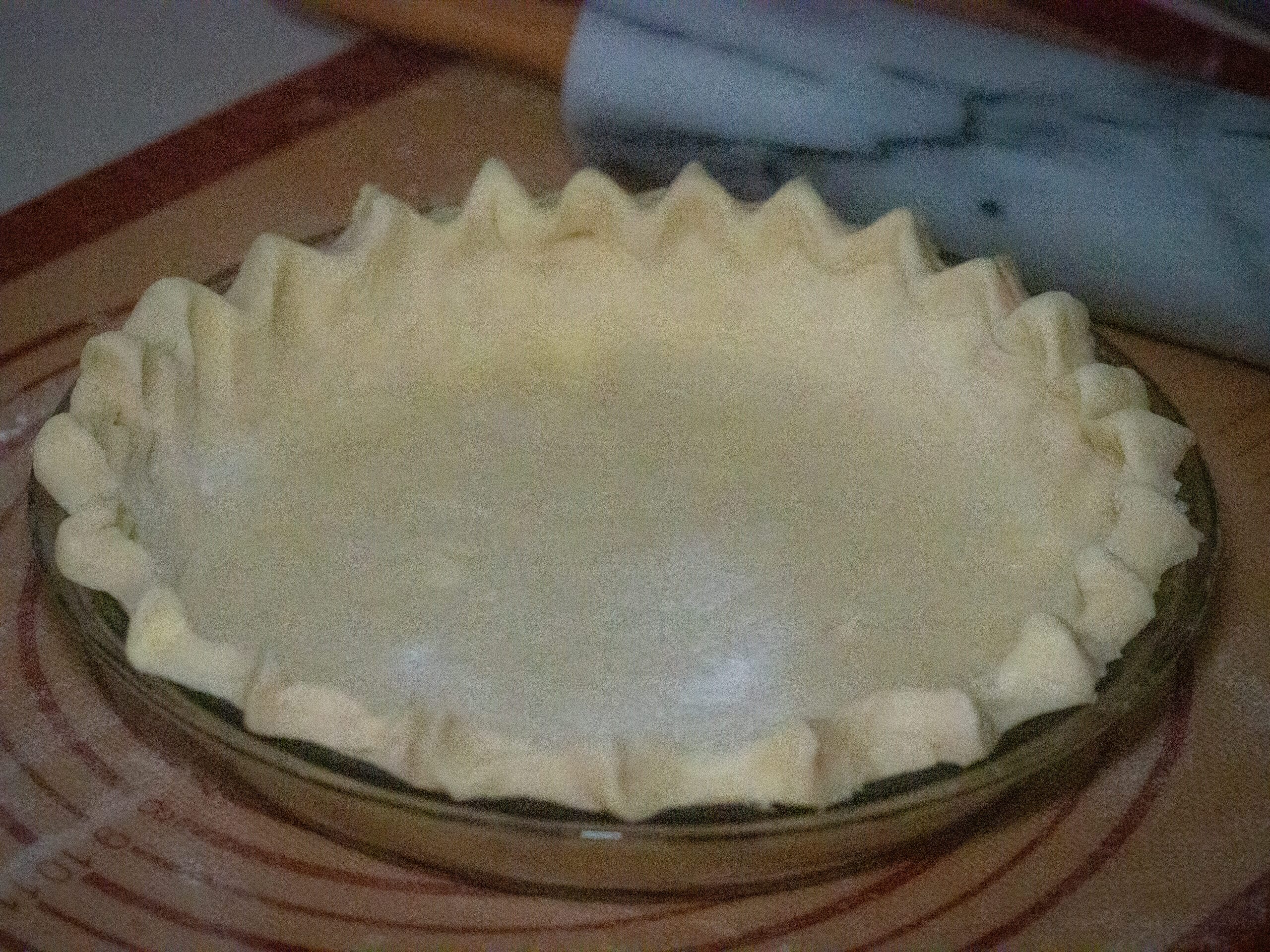 Pie dough in a glass baking dish on top of a brown silicon mat and marble rolling pin in the side