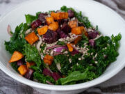 A bowl of chopped kale, roasted purple and orange sweet potatoes, thinly sliced red onion and sunflower seeds in a white serving bowl.
