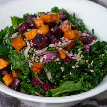 A bowl of chopped kale, roasted purple and orange sweet potatoes, thinly sliced red onion and sunflower seeds in a white serving bowl.