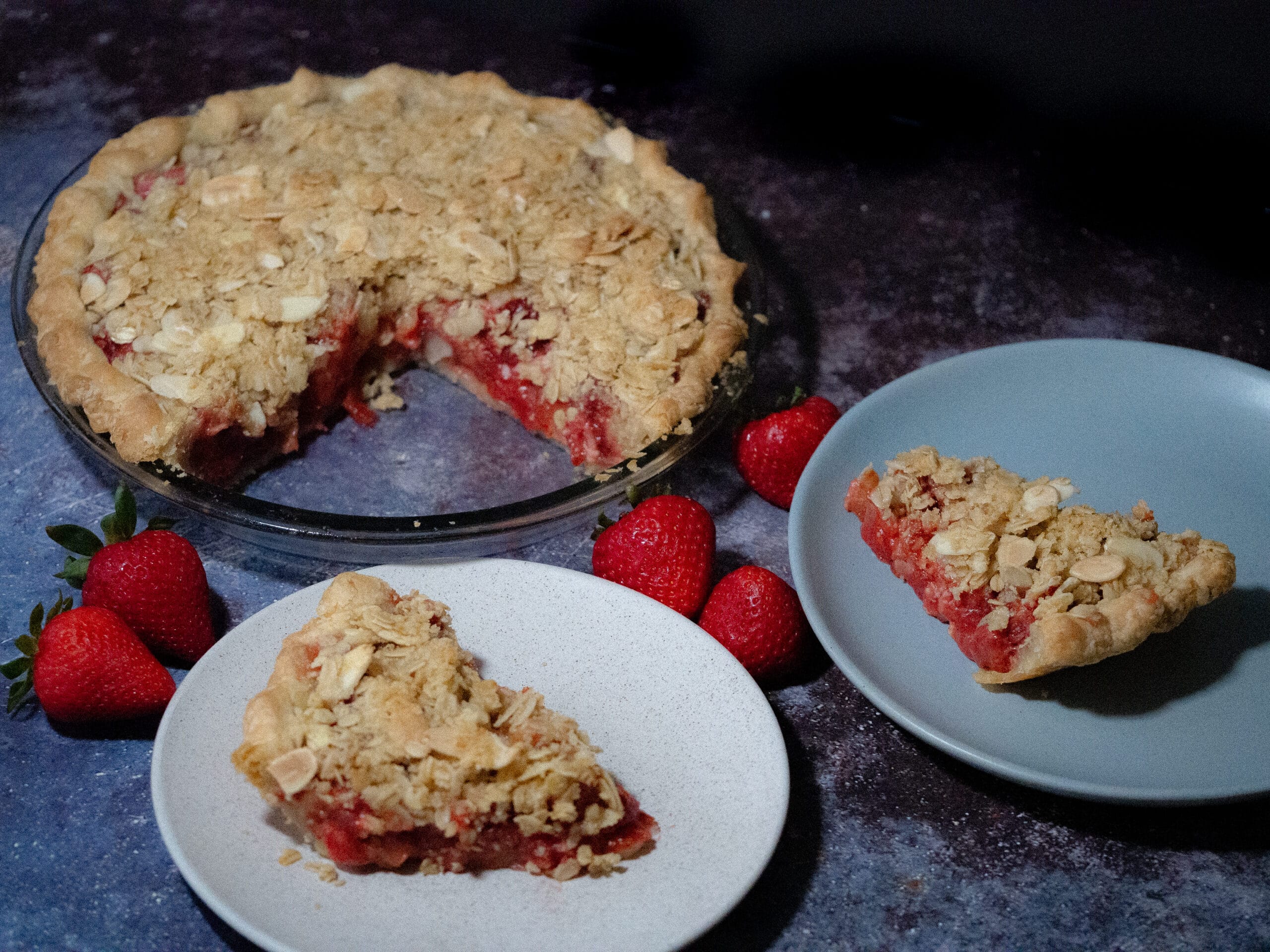 Sliced strawberry-rhubarb pie crisp with almond-oat streusel toppings on a dark grey patterned working space adorn with fresh strawberries.