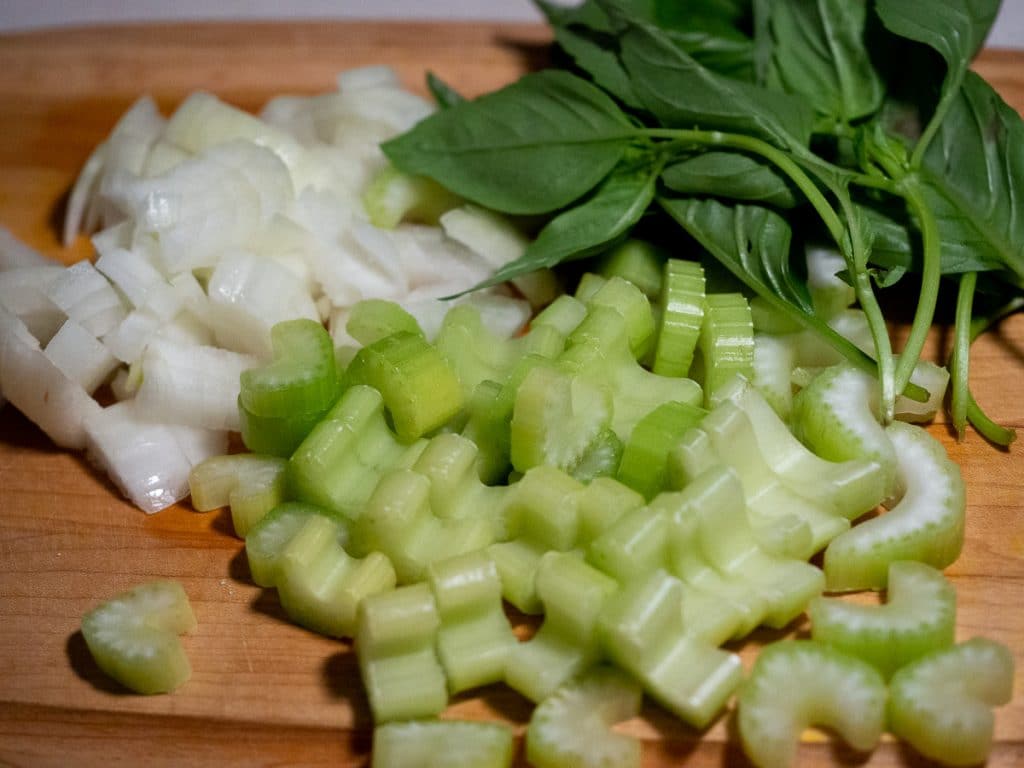 Diced onion and celery with stems of basil in the side on a wooden board