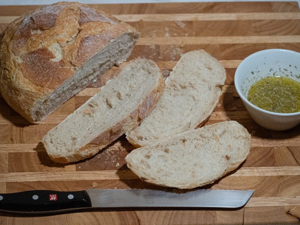 Whole and slice garlic bread on a wooden board with a bread knife at the front side and a bowl of garlic- olive oil and butter