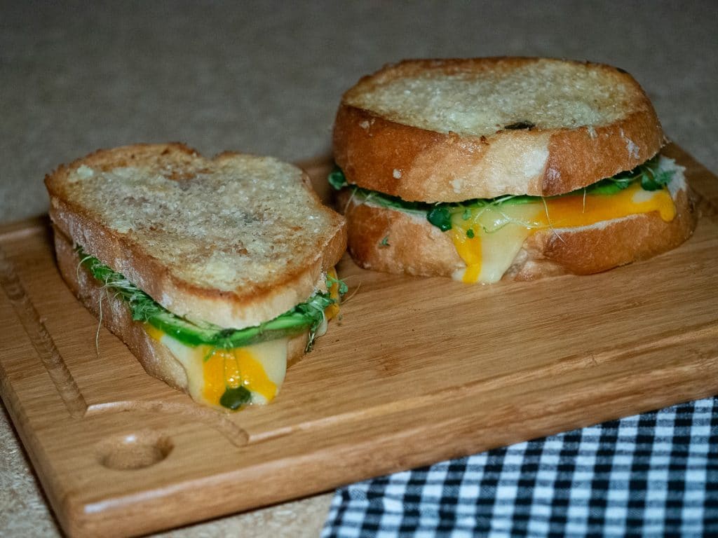 Two grilled cheese and avocado sandwich on a wooden board with a black and white linen on the side.