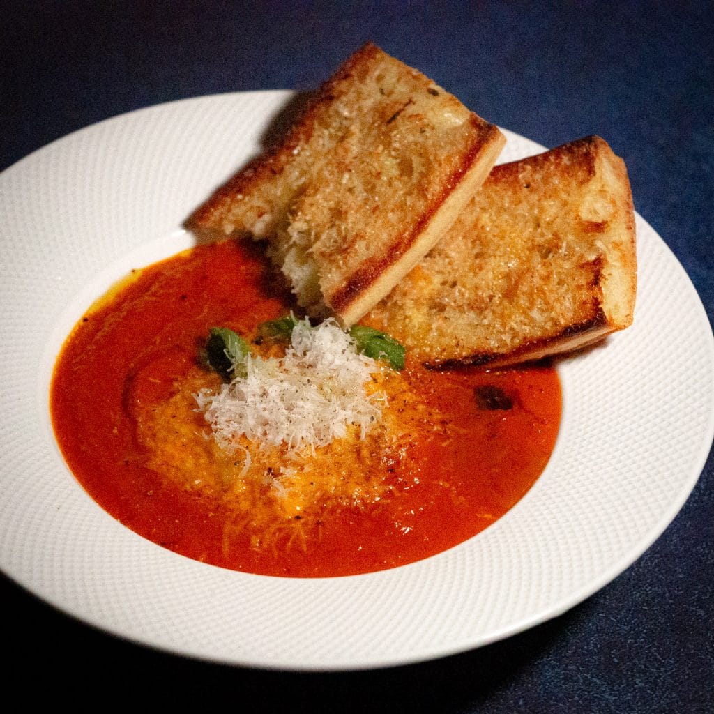Winter tomato soup in a white soup bowl with grated parmesan cheese and basil on top and garlic cheese bread on the side.