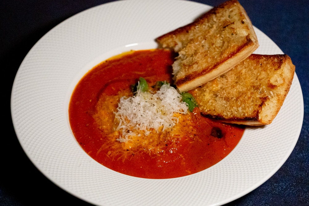 tomato soup with grated parmesan cheese on top garnished with a few basil leaf on a white soup bowl. Garlic-cheese read on the side.