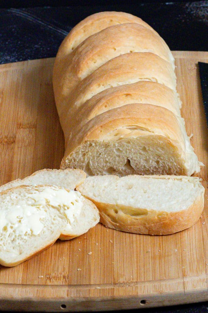 Golden brown whole French bread loaf ang a few slices slathered with butter on a wooden chopping board.