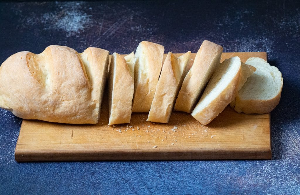 Slices of long loaf bread on a wooden chopping board on top of a dark blue surface.
