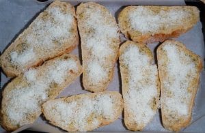 Slices of crusty bread layered with herb-garlic butter and top with grated parmesan cheese in a baking pan lined with parchment paper.