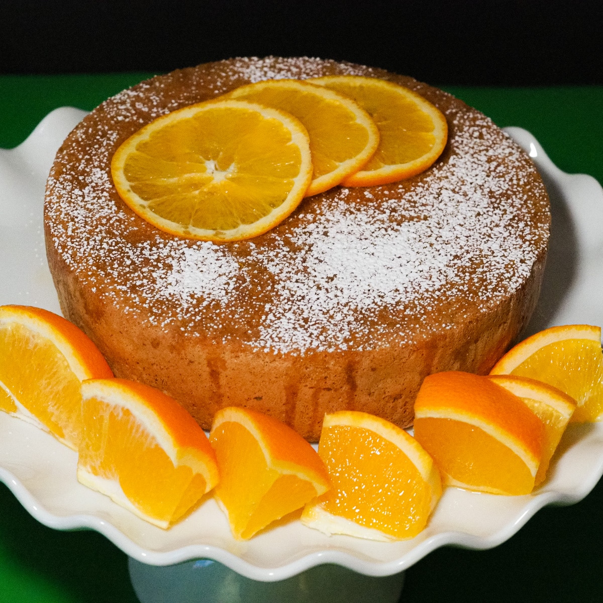 Alond-olive oil and orange cake dusted with icing sugar and 3 think slices of fresh orange place on top of a white ruffle cake stand and on the sides are thick slivers of fresh oranger.