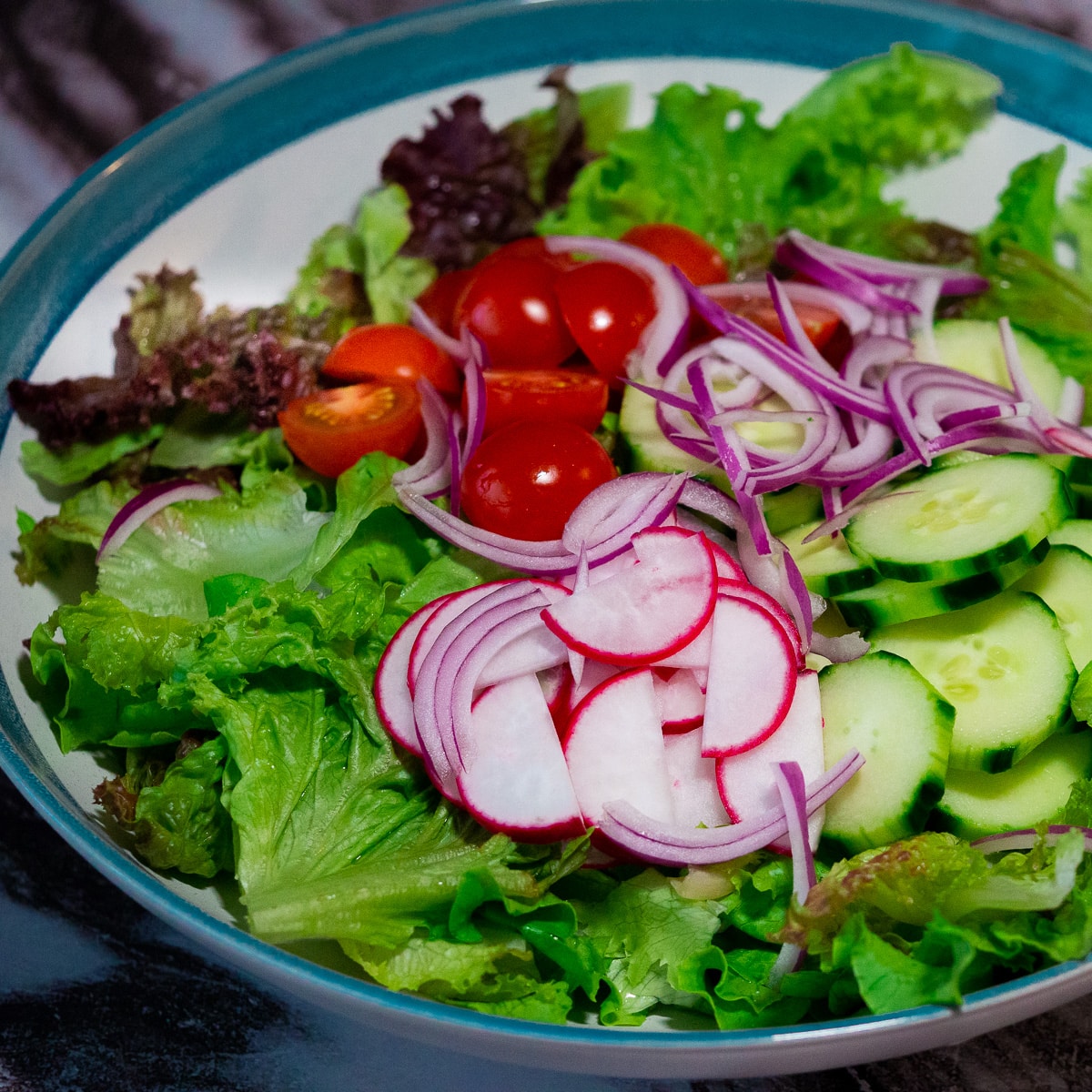 A mix of red and green oakleaf lettuce, cherry tomatoes, sliced raddish, sliced cucumber and sliced red onion in a a large serving bowl.