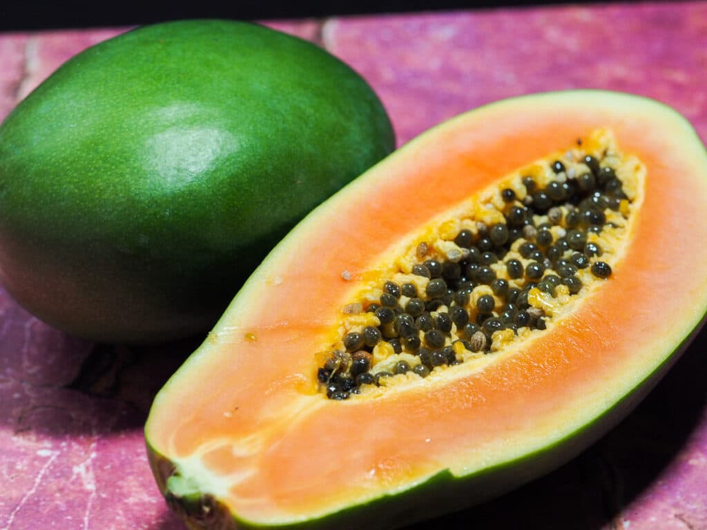 A whole green mango and half sliced ripe but firm papaya on a pink granite counter top.