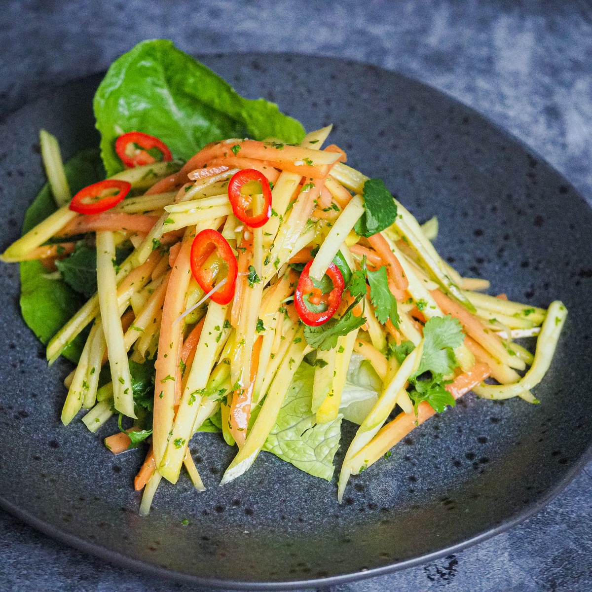 Thin slices of mango and papaya salad over romaine lettuce and garnished with slices of red chillies and cilantro in a grey plate with black dots placed on top of a grey-white counter top.
