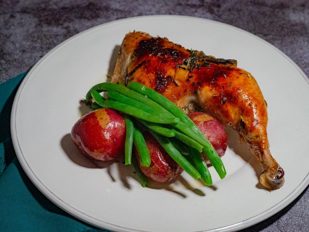 Roasted chicken quarter, roasted red potatoes and green beans on top in a white plate with green dish cloth on the left side.