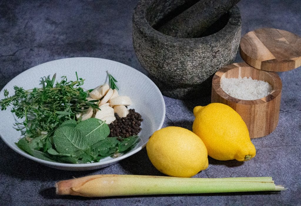 Mixed of fresh herbs, garlic and pepper in a white plate on a cement working space with 2 lemons, 1 lemon grass stalk, salt on a wooden container and pestle and mortar on the side.