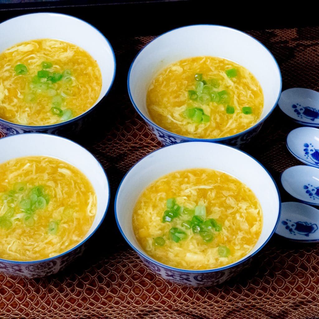 Yellow corn and egg drop soup in a four blue and white soup bowl with 4 ceramic soup spoon on the side placed on a brown leather serving tray.
