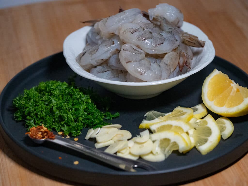 Raw shrimp in a white bowl over a black round plate with lemon slices, thinly sliced garlic, chopped parsley leaves and a teaspoon of chilli flakes.
