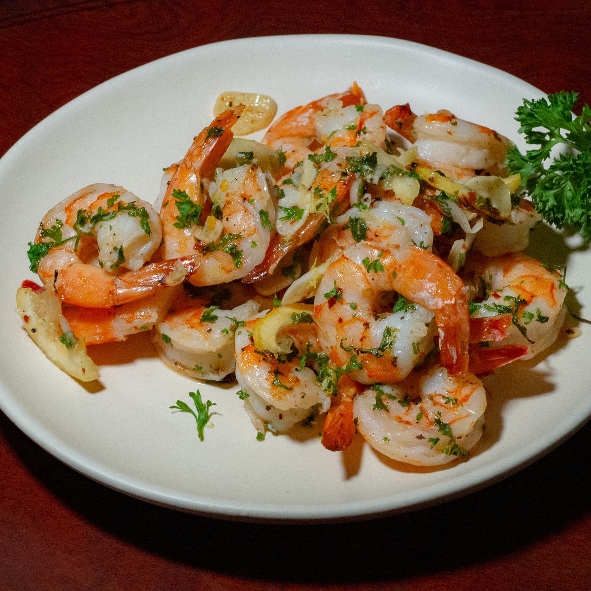 baked lemon garlic shrimp with thinly slice lemon, sliced garlic, chopped parsley and chilli flaces on a white plate placed on top of a brown surface.