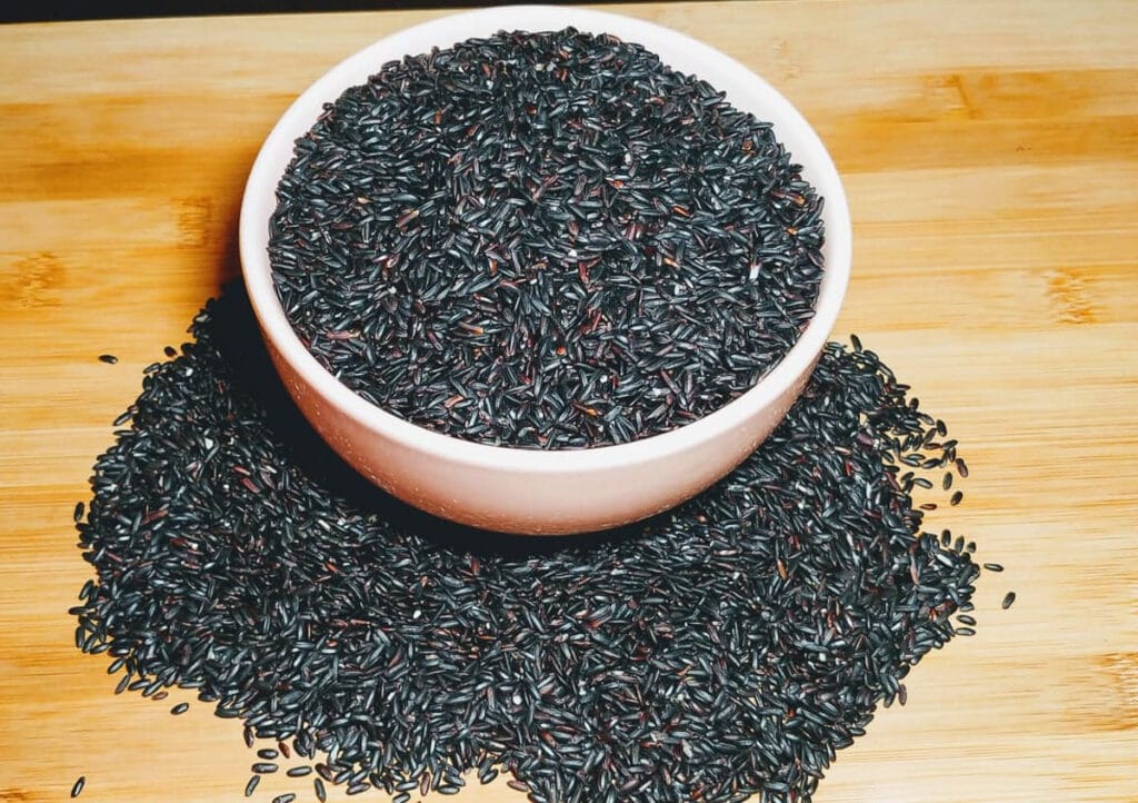 A bowl of black rice placed on a wooden boared with more black rice at the bottom