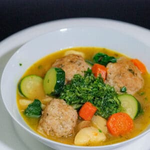 Three meat balls, chopped potatoes, sliced zucchini, chopped potatoes and broth with green sauce at the middle in a white bowl