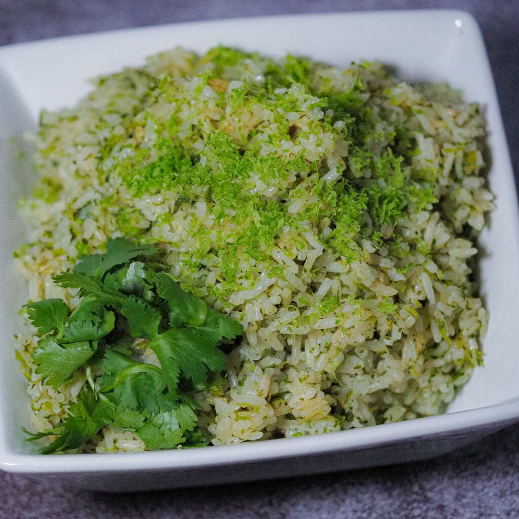 Cooke herby rice in a white square bowl topped with lime zest and garnished with fresh cilantro at the front left corner,