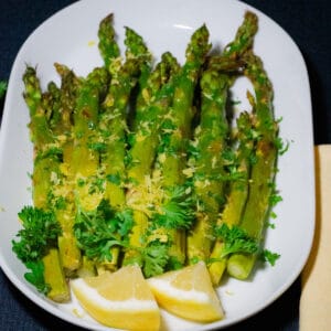 Perfectly lemon roasted asparagus arranged in an oval plated garnished with parsley and lemon zest and 2 lemon slices on the side
