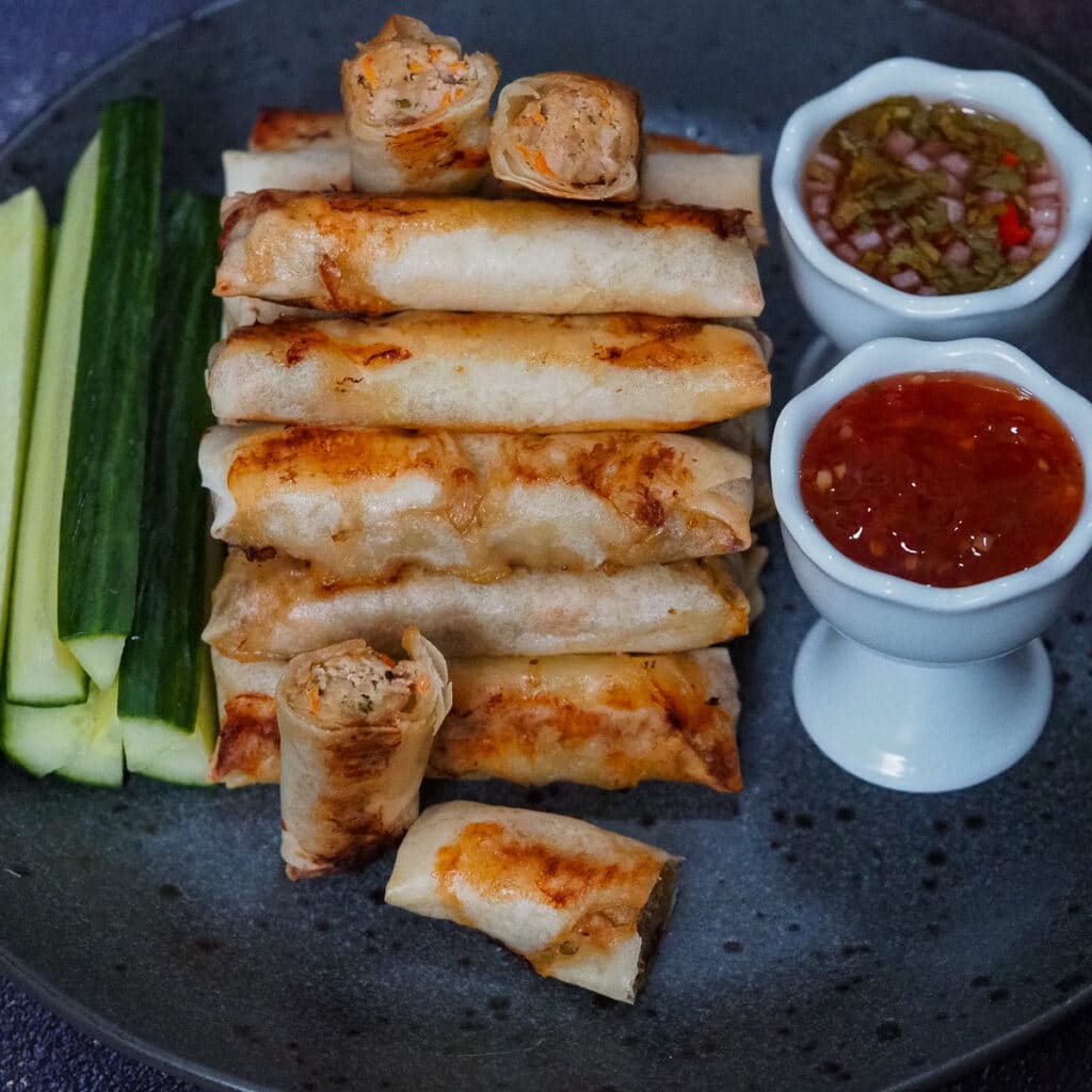 Baked lumpiang shanghai piled on a black plate with slices of cucumber and chilli sauce on the side