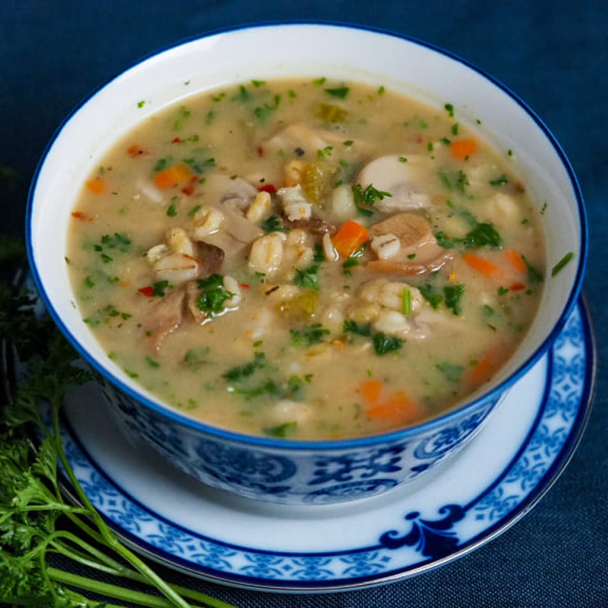 Wheat, barley-oat blend soup with sliced mushroom, diced carrots and chopped parsley in a bowl with blue rim placed on a blue rimmed plate on top of a dark blue counter top adorn with parsley on the left side.