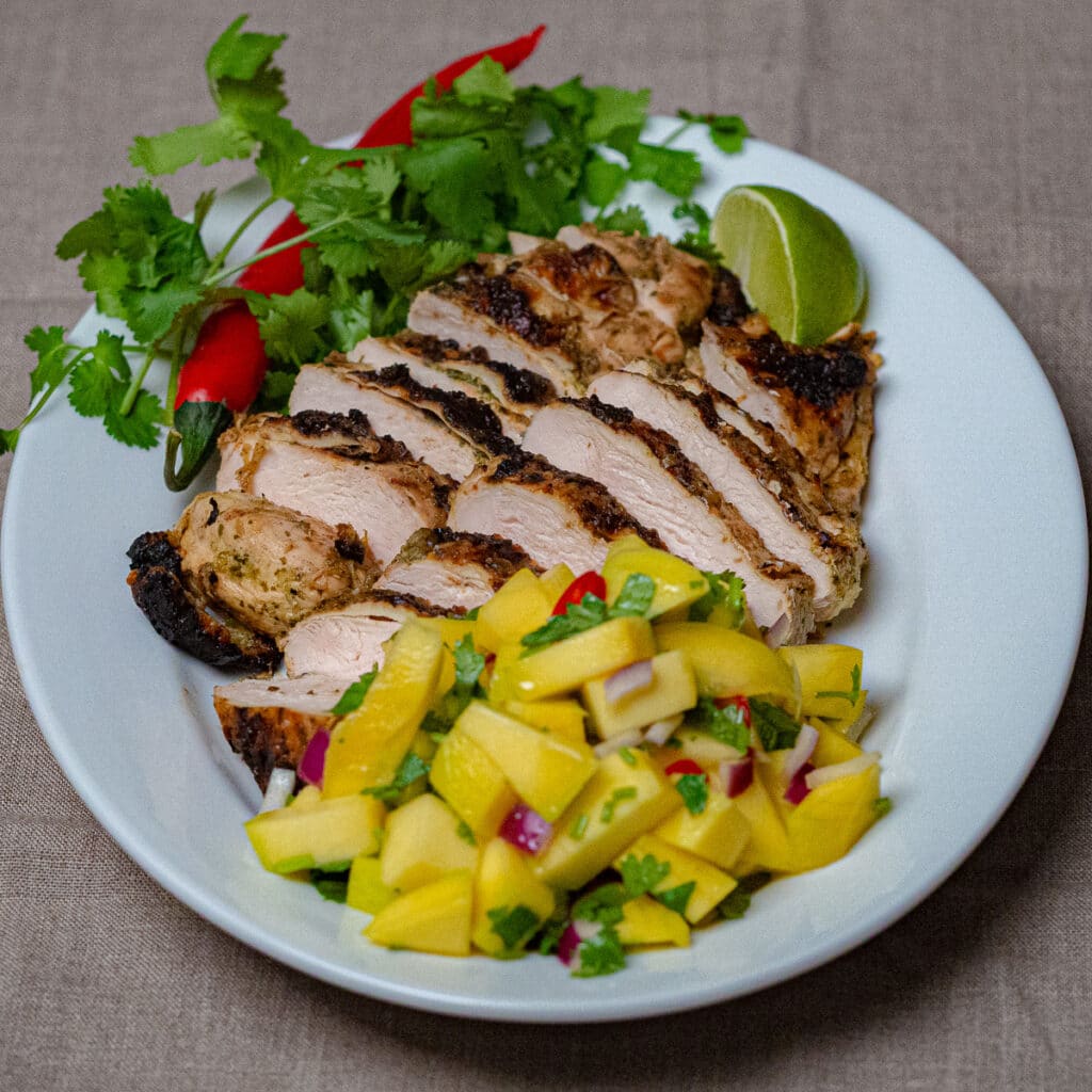 Sliced grilled chicken breast and mango salad in a white oval serving plate grnished with stems of cilantro, red chilli and a slice of lime