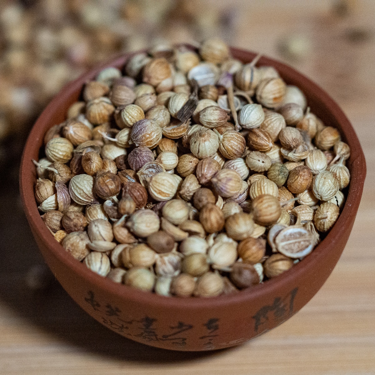 Coriander seeds in a dark brown pinch bowl on a wooden surface.