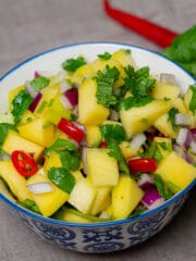Diced mango salad chopped chilli, red onion and cilantro in a white and blue rimmed bowl on a beige surface adorn with cilantro and red chilli on the side.