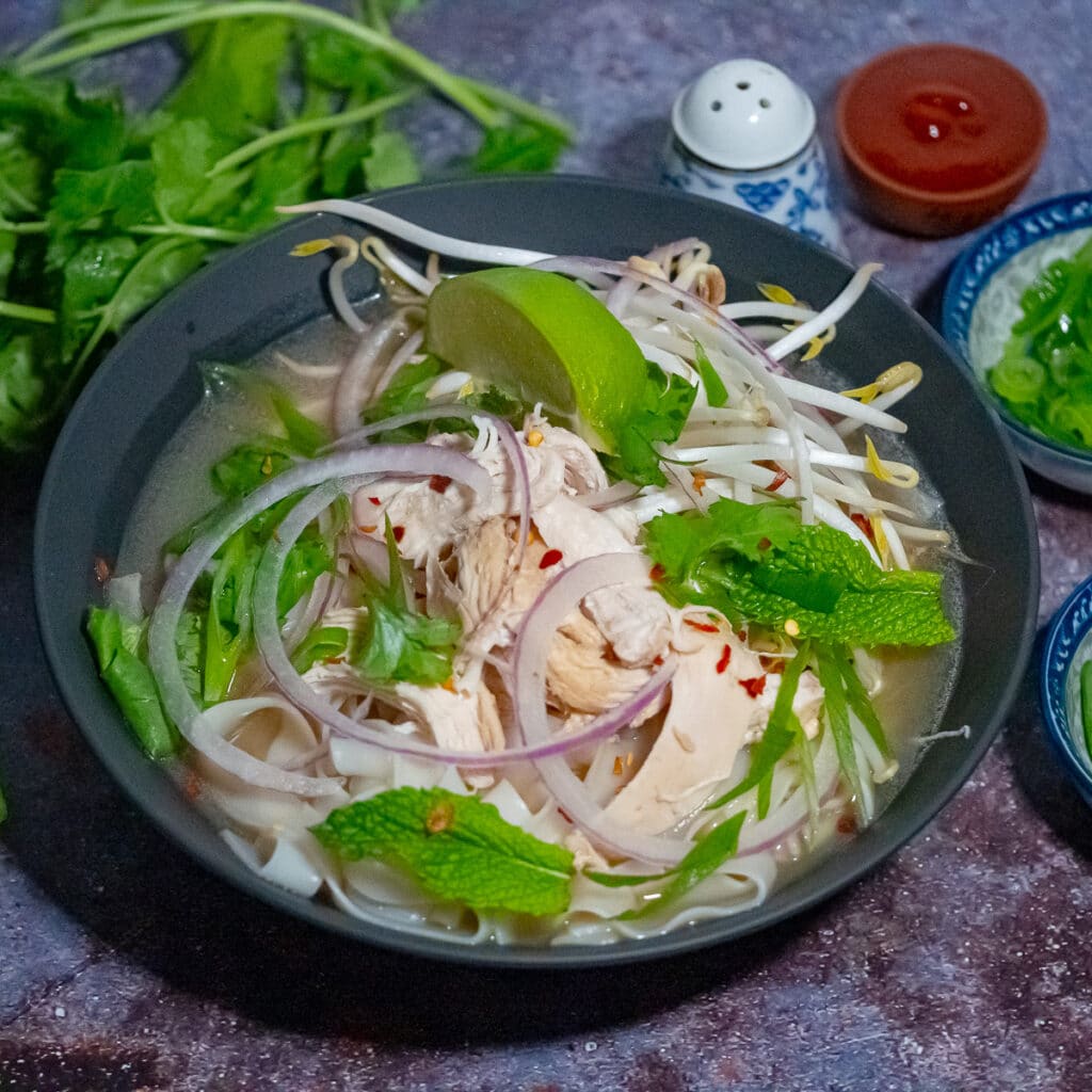 Chicken pho soup in a black bowl top with shredded chicken, green herbs, slice of lime, sliced red onion, and sprouts with stems of cilantro, salt shaker, chilli sauce, and sliced herbs on the side.