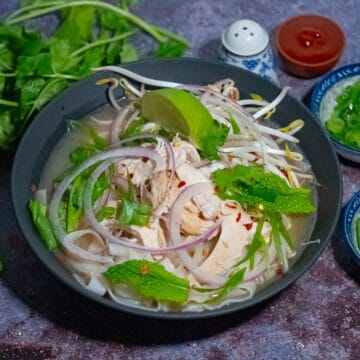 Chicken pho soup in a black bowl top with shredded chicken, green herbs, slice of lime, sliced red onion, and sprouts with stems of cilantro, salt shaker, chilli sauce, and sliced herbs on the side.