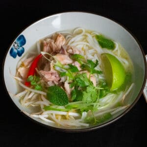 Chicken pho soup in a bowl with rice noodles, shredded chicken, chili, slice of lime and green herbs on top