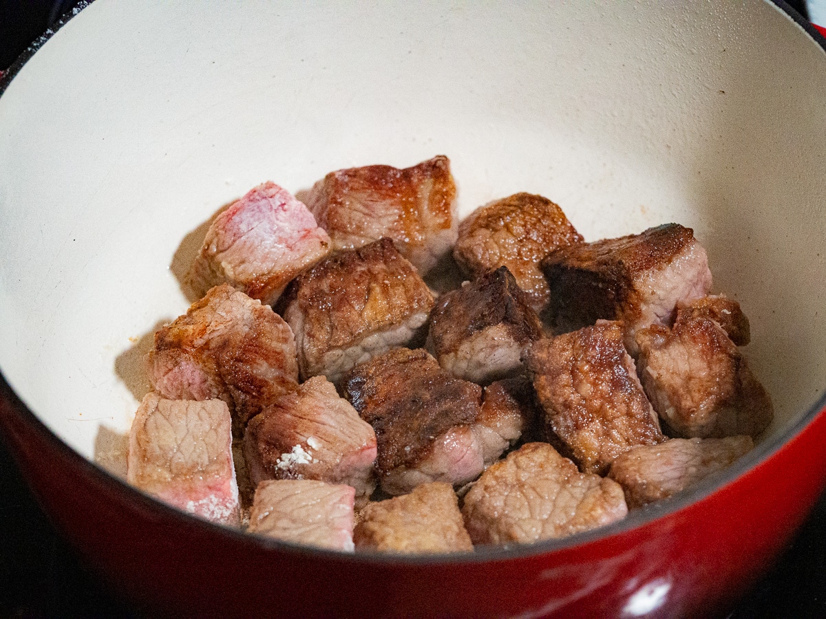 Searing chunky slice of beef to deep brown in a white and red pot.