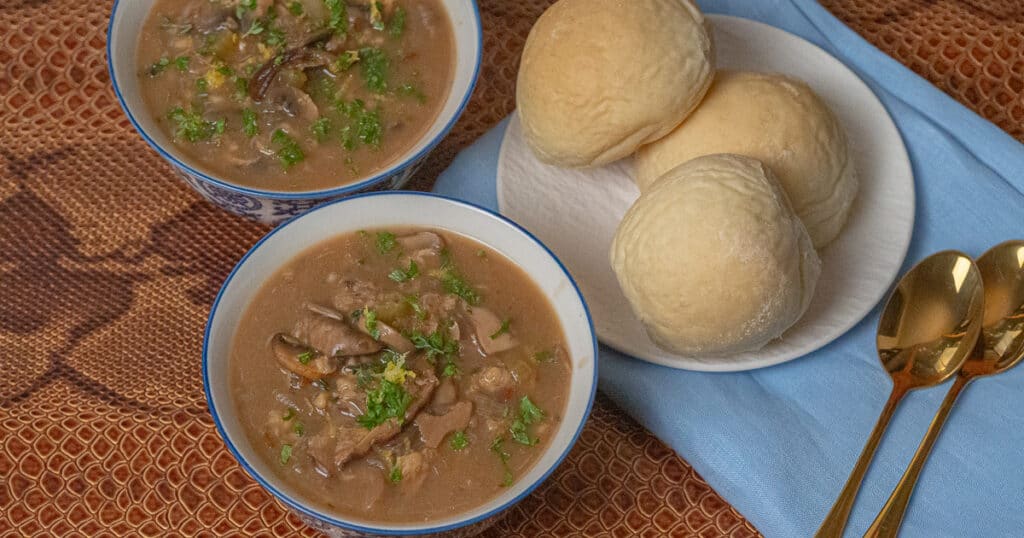 2 bowls of mushroom soup, 3 buns in a white plate over blue cloth and 2 gold soup spoon placed in a brown leather serving tray.