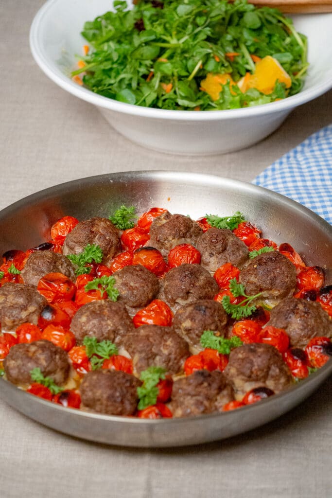 Oven roasted beef meatballs and cherryt omatoes in an oven proof skillet garnished with chopped parsley and a bowl of salad on the side