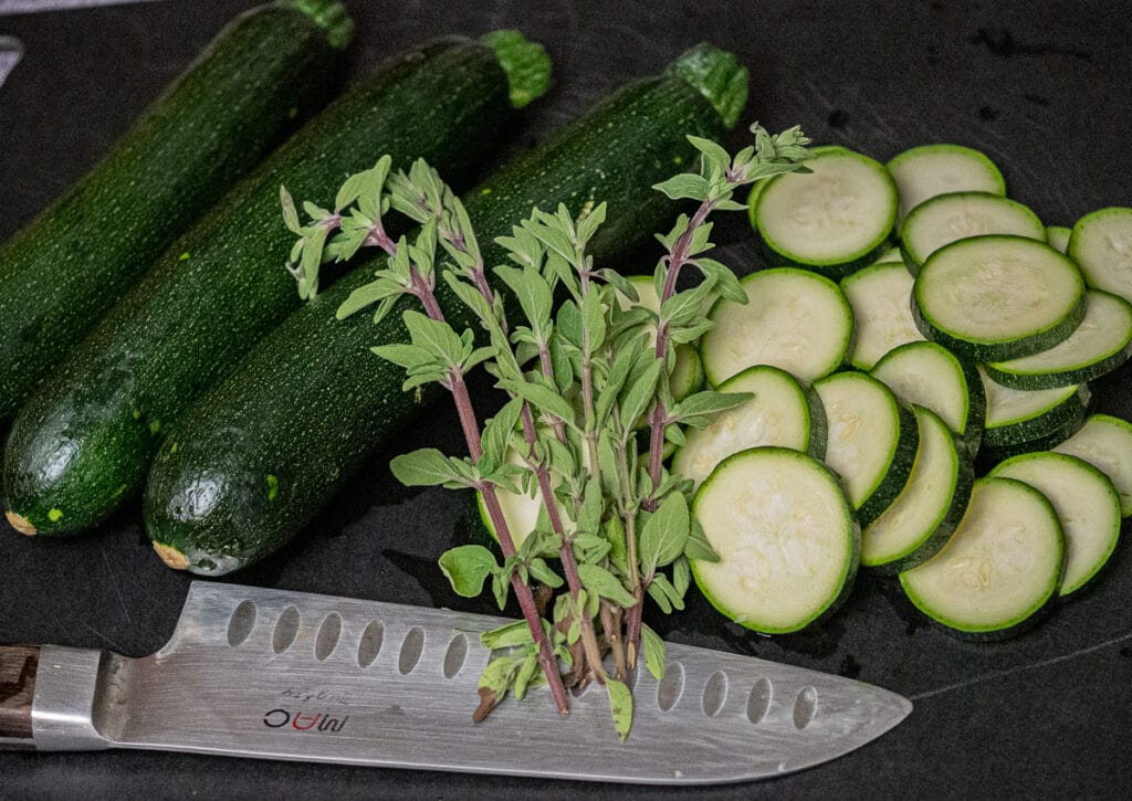 3 pieces of zucchini, steems of oregano, sliced zucchini and a knife on the side on top of a black chopping board.