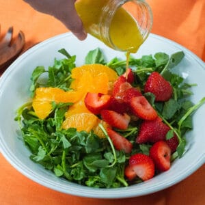 Watercress, oranges and strawberries in a white serving bowl while drizzling a lemon-garlic vinaigrette