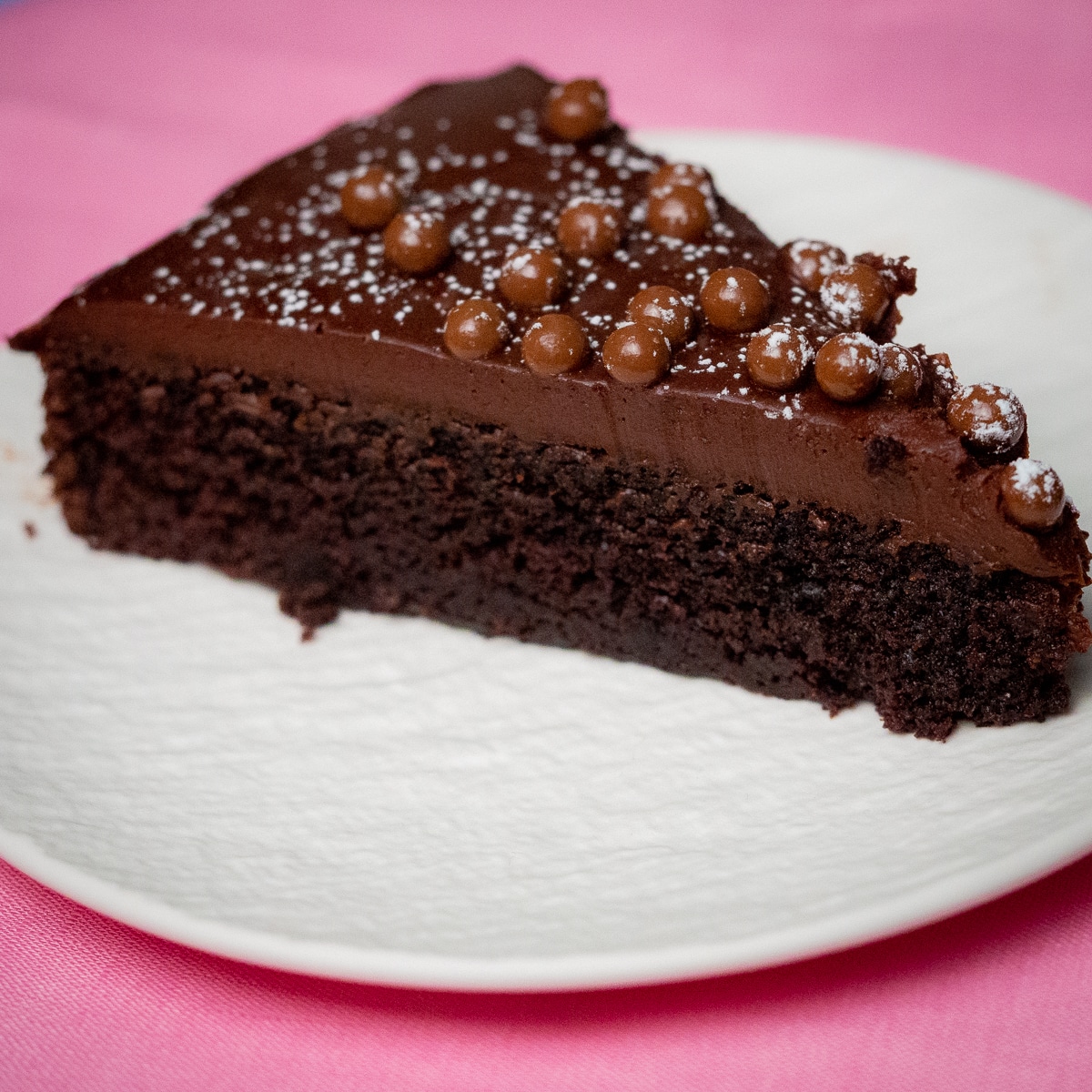 A slice of chocolate fudge cake topped with chocolate pearls and a dust of icing sugar in a white dessert plate placed on top of a pink surface