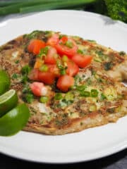 Eggpant omelette wth diced tomatoes and green onions on top placed on a white plate.. Adorn with lime wedges and fresh parsley on the side.