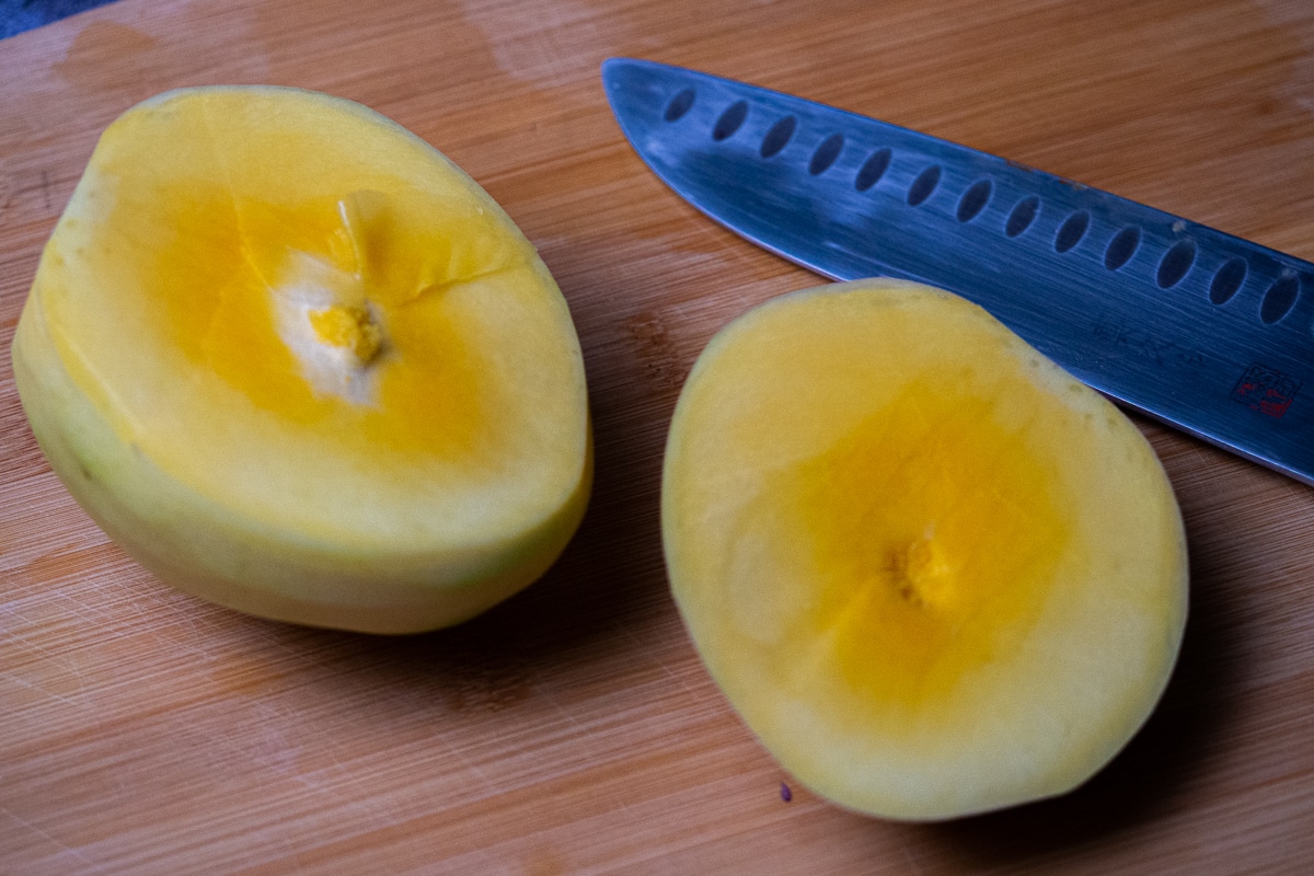 Peeled and cut in half green mango placed on top of a wooden board and a knife on the side