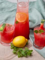 Two glasses of watermelon lemonade garnished with slices or watermelon and 1 jug Placed on a brown linen covered workspace adorn with green linen, one lemon, mint leaves and two glass straw.