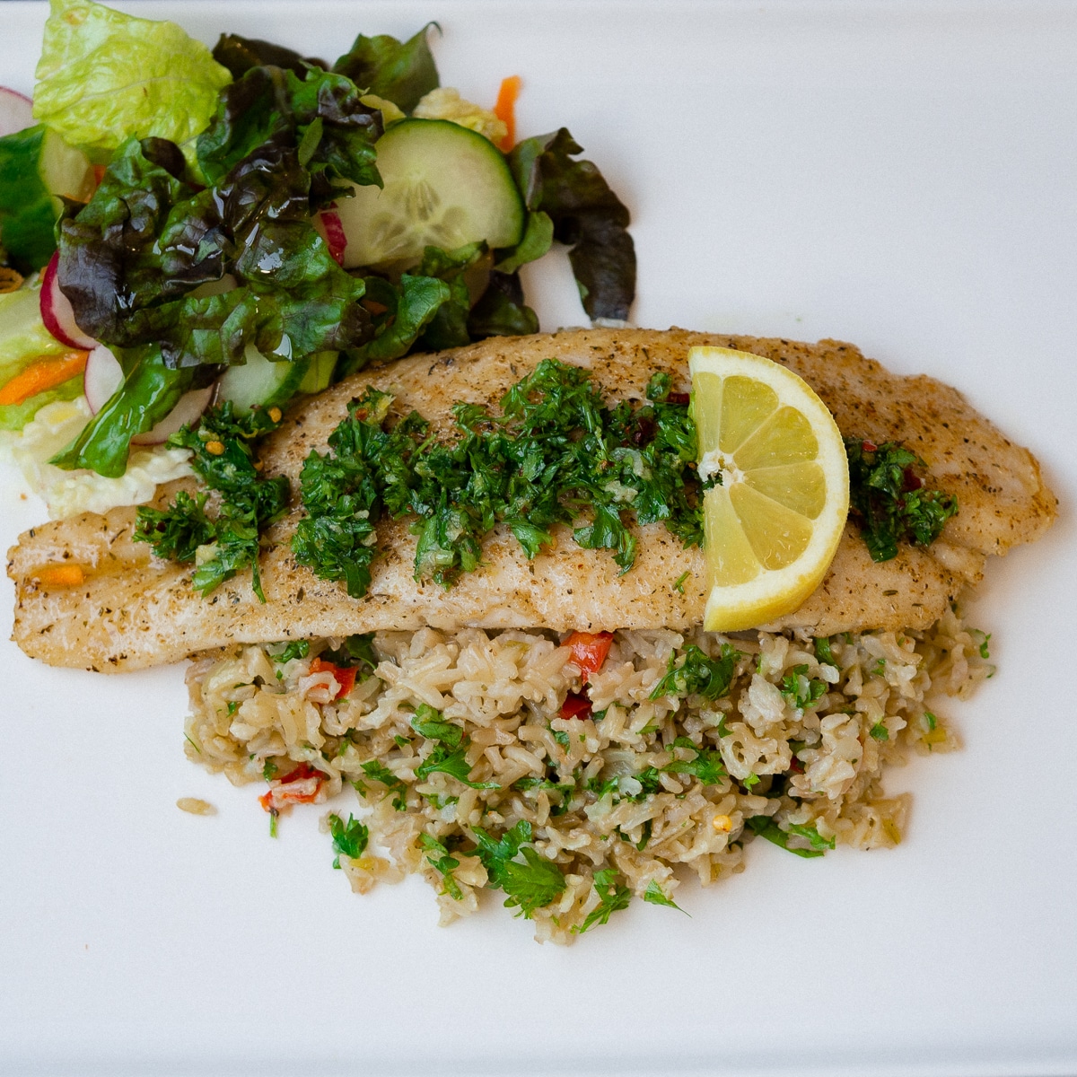 Brown basmati rice pilaf top with fish fillet gremolata and chopped salad on the side on a white plate.