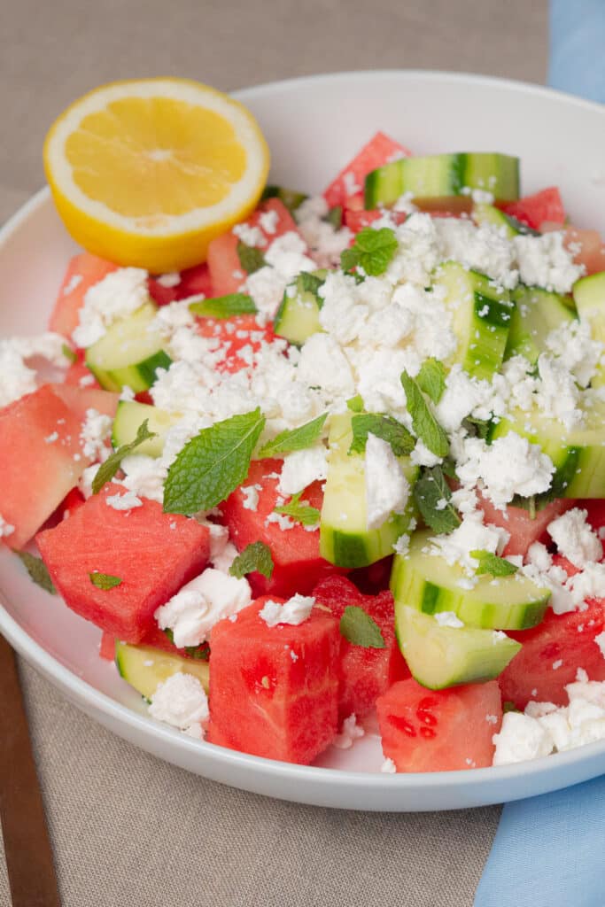 Cubed watermelon and chunky slices of cucumber on a white large bowl toppped with crumbled feta cheese, scattered mint leaves and a half sliced lemon.